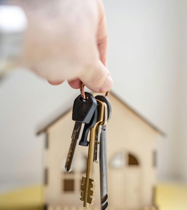Close-up of a hand holding keys with a miniature wooden house in the background, symbolizing real estate investment.
